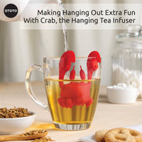 Cute Tea Infuser by OTOTO shaped like a red crab hanging on a glass mug, steeping loose leaf tea. A fun tea accessory for kitchen or gift use.