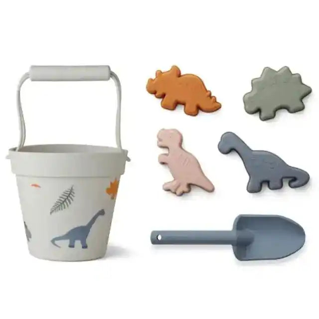 Dinosaur-themed beach toy set with a white bucket featuring dinosaur illustrations, a matching shovel, and dino-shaped sand molds for kids.