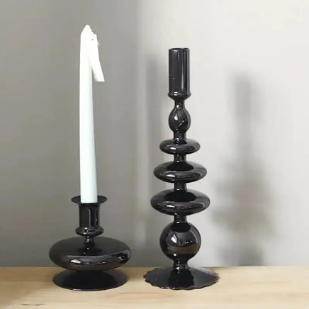 Two black Rue glass candlesticks, one holding a white taper candle, displayed on a wooden surface with a neutral background.