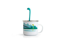 Cute Tea Infuser by OTOTO shaped like a sea monster, placed in a white enamel mug with a lake and forest design. Fun tea accessory for loose leaf tea.