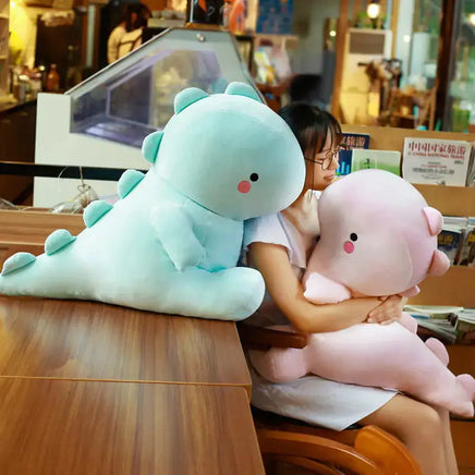 Two large dinosaur plush toys, one blue and one pink, being hugged by a woman in a cozy café setting.