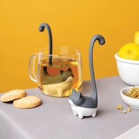 Cute Tea Infuser by OTOTO shaped like a cat with a long tail, steeping loose leaf tea in a clear mug on a table with cookies and lemons nearby.