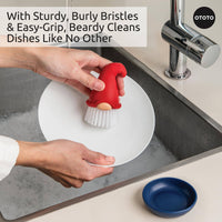 Beardy Gnome Dish Brush by Ototo in use, scrubbing a plate with sturdy bristles and ergonomic grip, includes a blue drip tray.