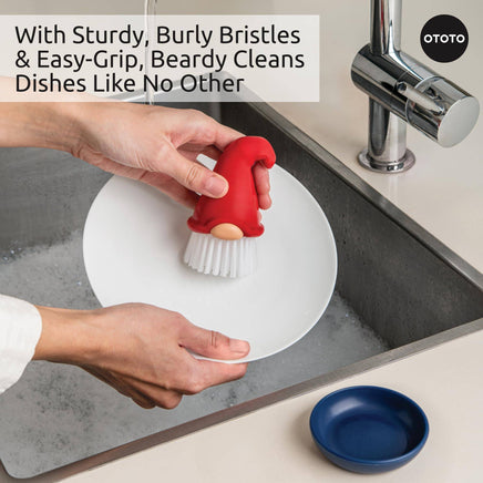 Beardy Gnome Dish Brush by Ototo in use, scrubbing a plate with sturdy bristles and ergonomic grip, includes a blue drip tray.