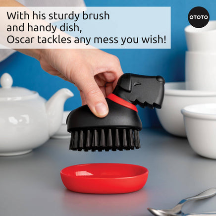 Oscar Dish Brush by Ototo in action, scrubbing a plate with sturdy bristles and ergonomic grip, includes a red drip tray.