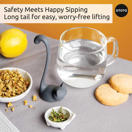 Cute Tea Infuser by OTOTO shaped like a cat with a long tail, ideal for loose leaf tea, shown beside a glass mug, herbs, and cookies on a table.