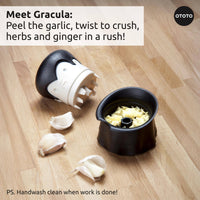 OTOTO Gracula garlic crusher with vampire design on wooden counter, shown crushing garlic cloves. Fun Halloween kitchen gadget for easy prep.