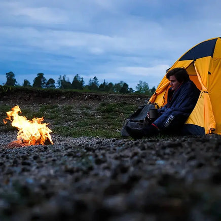 Camper wrapped in a portable sleeping bag near a campfire beside a yellow tent. Ideal for outdoor adventures, ensuring warmth and comfort.