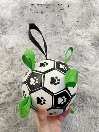 Hand holding a black and white dog football toy with paw prints and green straps on a gray carpet.