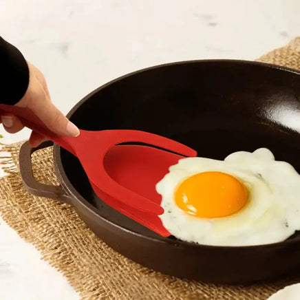 Red 2-in-1 kitchen gadget flipping a fried egg in a pan, showcasing its spatula and fork functionality for versatile cooking.