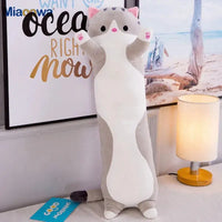 Gray Cute Cat Pillow Plush Toy standing upright on a table beside a lamp, ideal for hugging or as a decorative piece.