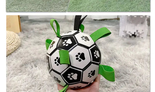 Black and white dog football toy with paw print design and green straps, placed on a gray carpet.