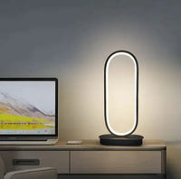 Oval LED table lamp with a sleek black base, placed on a desk beside a laptop, emitting a soft white glow.