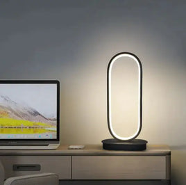 Oval LED table lamp with a sleek black base, placed on a desk beside a laptop, emitting a soft white glow.