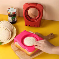 Pink stainless steel sandwich cut mold in use, creating round toast pockets on a wooden board with bread and jam in the background.