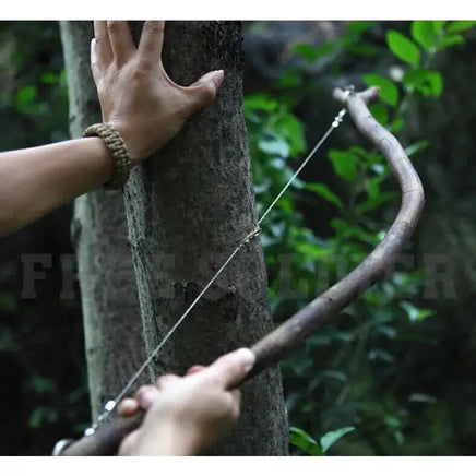 Person using a steel wire saw attached to a wooden stick to cut a tree branch, demonstrating its utility in survival situations.