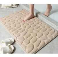 Beige super absorbent shower room floor mat with a pebble-like texture, placed near a shower area with slippers nearby.