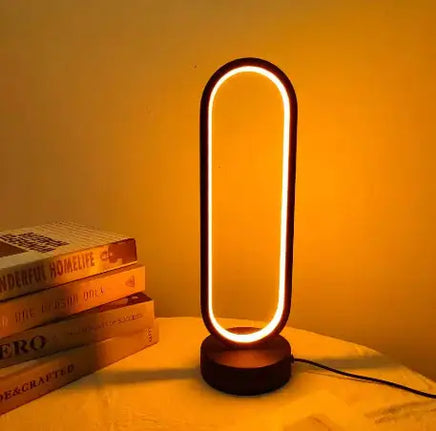 Oval LED table lamp with a black base, emitting a warm amber glow, placed on a table beside stacked books.