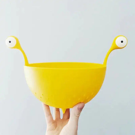 Hand holding OTOTO Draining Basket for Kitchen, highlighting its compact size, bright yellow color, and playful monster-themed handles.