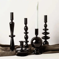 Collection of Rue glass candlesticks in sleek black designs, displayed with a white taper candle on a soft brown fabric.