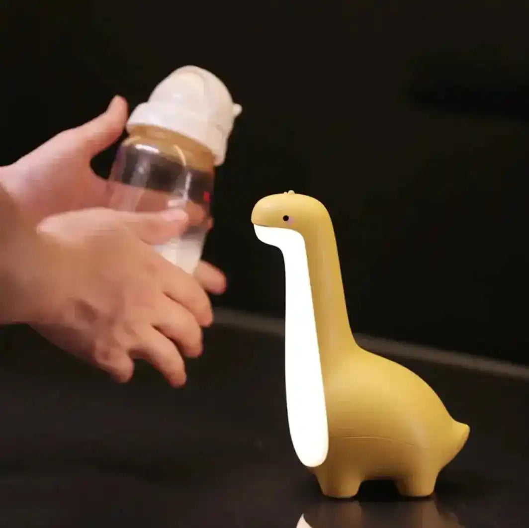 Yellow dinosaur night light glowing softly while a hand holds a baby bottle, highlighting its practicality for nighttime feed