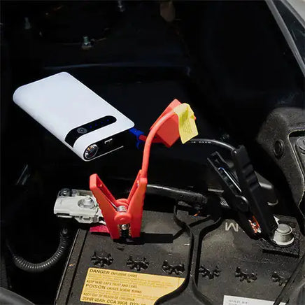 Car jump starter connected to a vehicle battery with clamps, demonstrating its use in emergency vehicle repair situations.