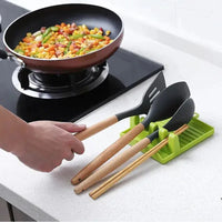 Green spatula drain rack gadget holding spatulas and chopsticks on a kitchen counter next to a cooking pan.