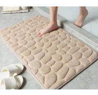 Beige super absorbent shower room floor mat with a pebble-like texture, placed near a shower area with slippers nearby.