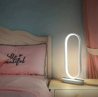 Oval LED table lamp with a white base, placed on a bedside table, providing soft white light in a cozy bedroom setting.